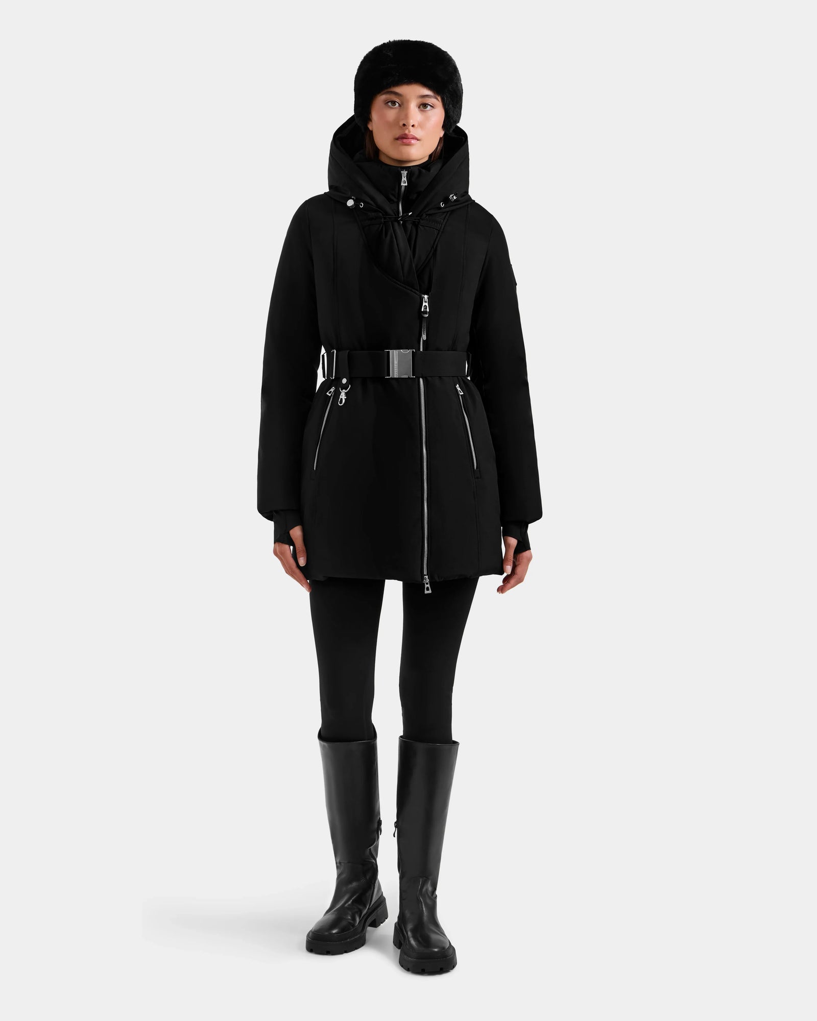 A person stands facing forward, wearing the Rudsak ISELLE - L125547 Black women’s down parka with a belt, black pants, knee-high boots, and a black fur hat against a light gray background.