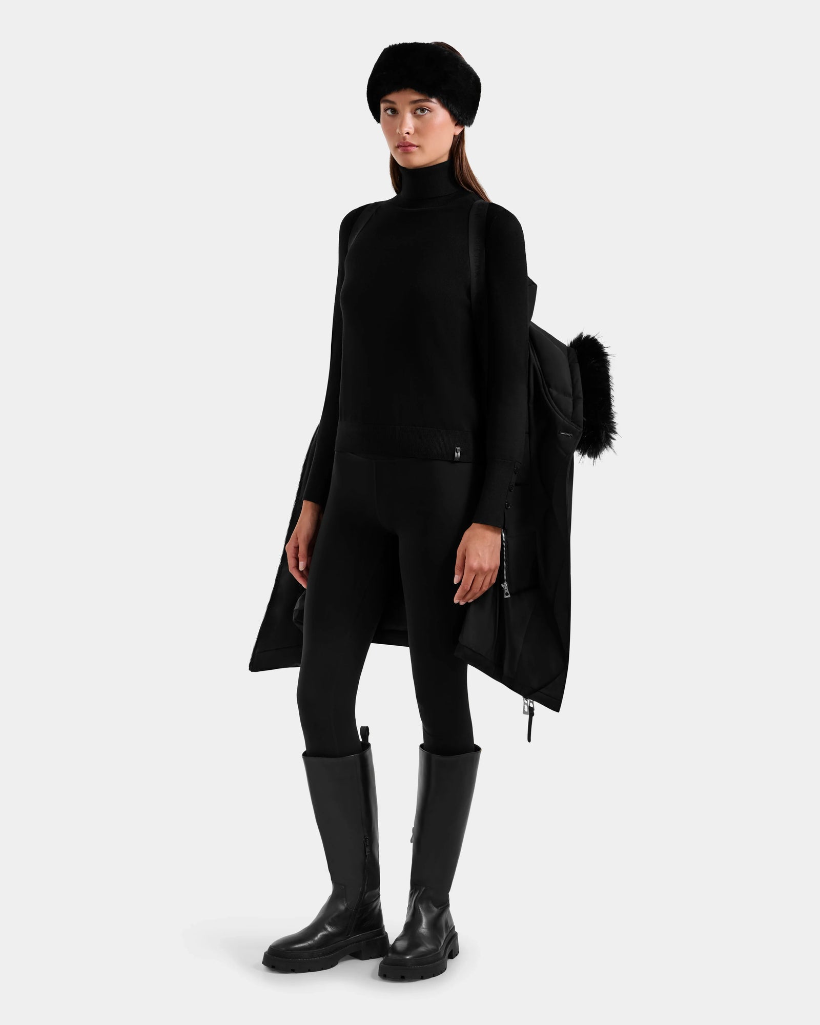 A woman faces forward, wearing the Rudsak ISELLE - L125547 Black winter coat over a turtleneck, leggings, tall boots, and a fur hat against a plain white background.