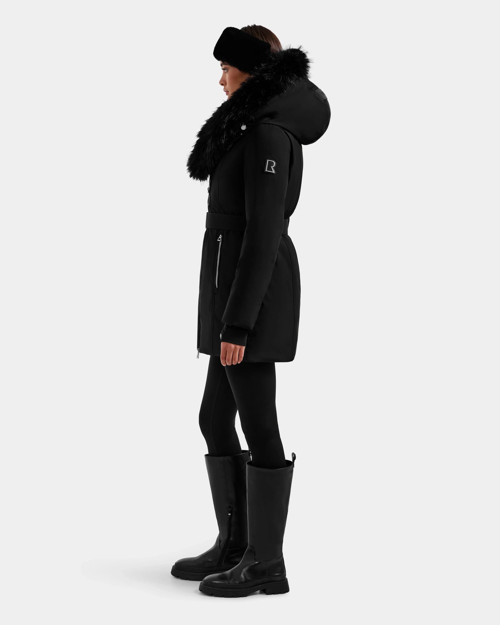 A person in profile models the Rudsak ISELLE - L125547 Black women’s down parka with a fur-trimmed hood, styled with black leggings, tall black boots, and a black headband against a plain light background.