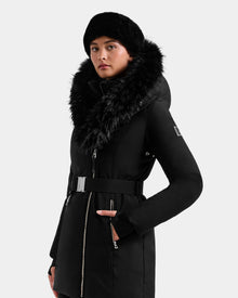 A person wearing the Rudsak ISELLE - L125547 Black belted women’s down parka with a large faux fur-trimmed hood and a black fur hat, standing against a plain light background.