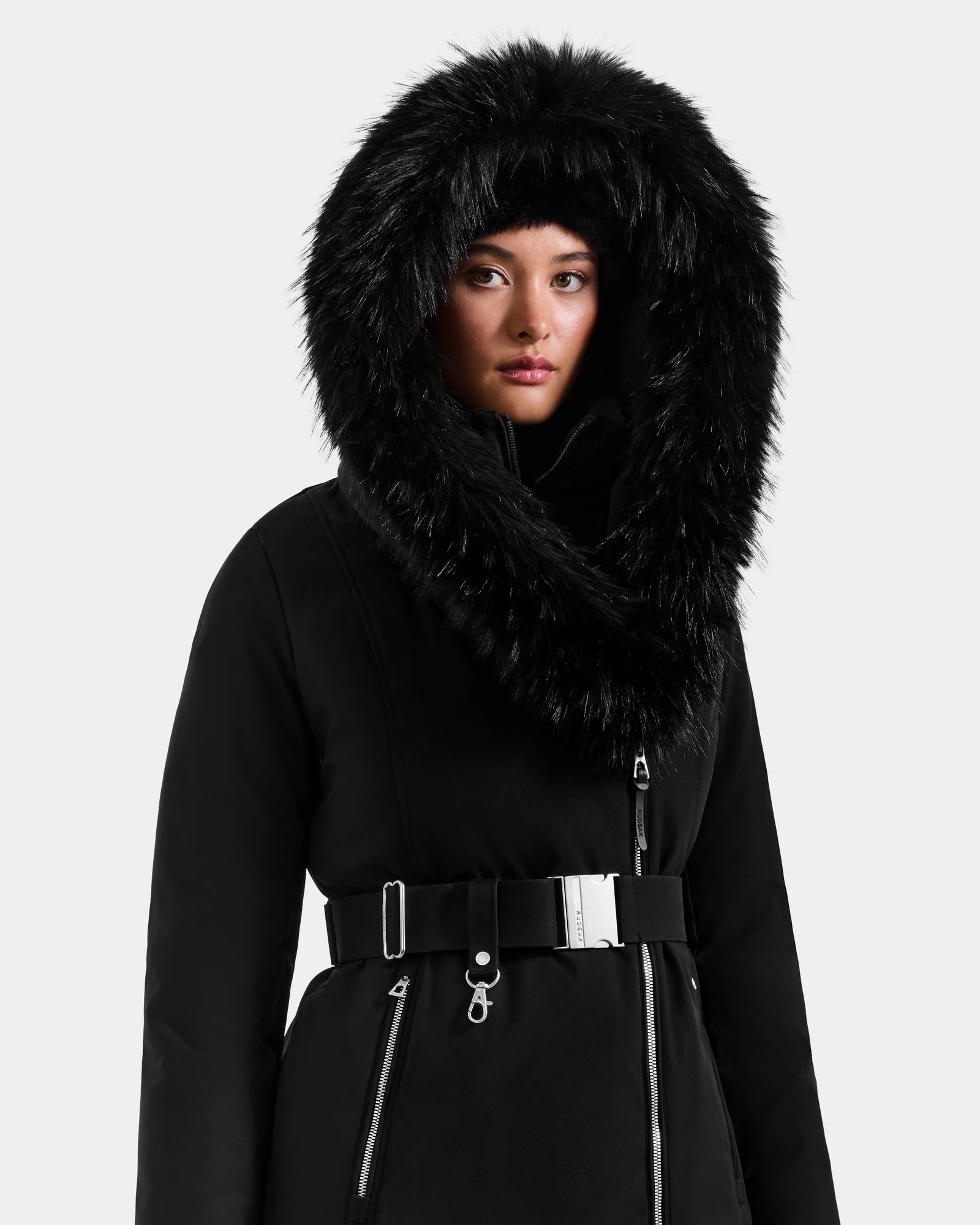 A person wearing the Rudsak ISELLE - L125547 Black women’s down parka with a large fur-trimmed hood and belted waist stands against a plain light background, looking forward.