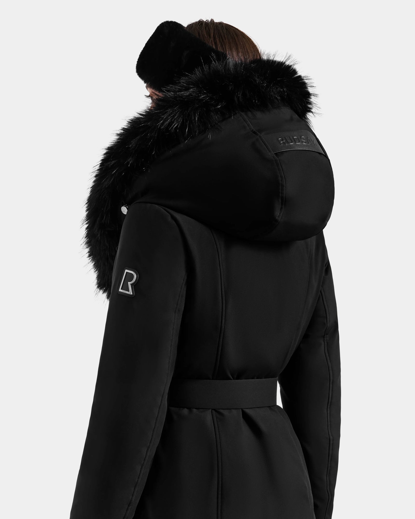 A person is shown from the back against a plain white background, wearing the Rudsak ISELLE - L125547 Black women’s belted down parka with a large furry hood and a matching black ear warmer.