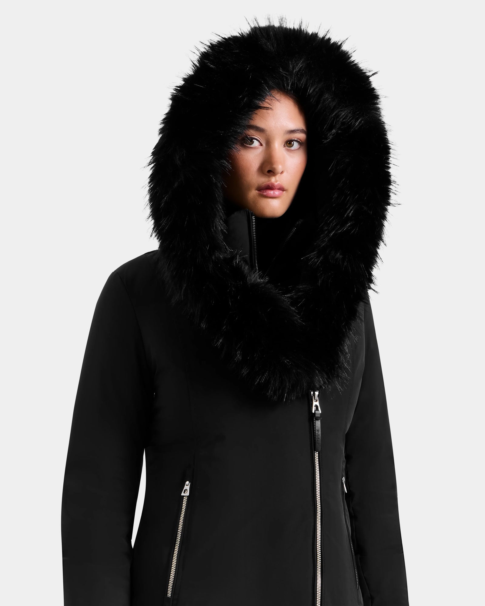 A woman wearing the Rudsak JANIS - L125558 Black parka with a large, fluffy black fur hood looks forward with a neutral expression against a plain light background—a stylish cold-weather layer.