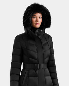A person wearing the Rudsak JULIETA M-L - L125559 Black belted maxi down puffer coat with a large faux fur hood and weather-resistant shell poses against a light gray background.