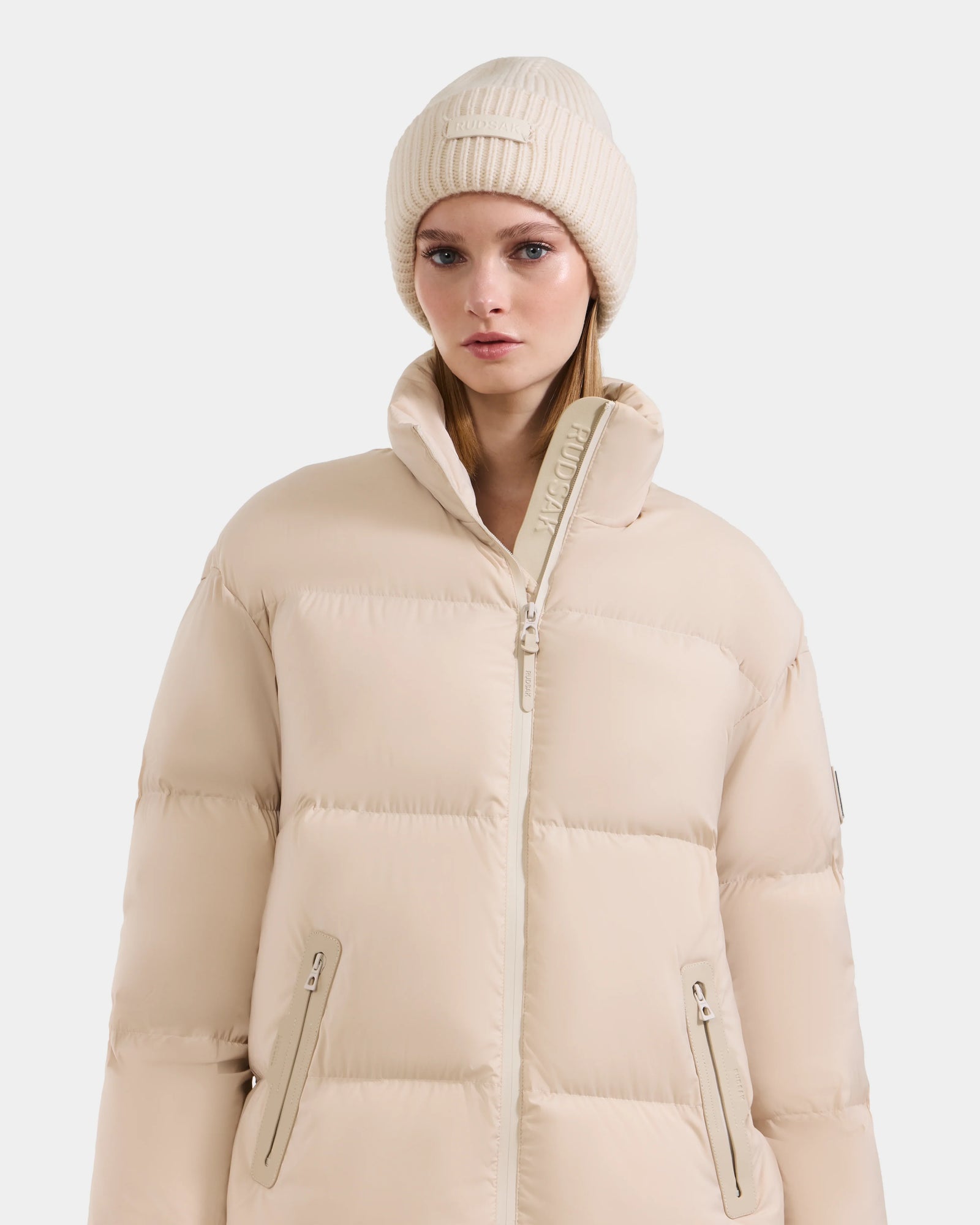 A woman wears the Rudsak KARLEE L - 8125682 Vanilla jacket with zippered pockets, vegan leather trims, and a high collar, paired with a matching knit beanie, posing against a plain white background.