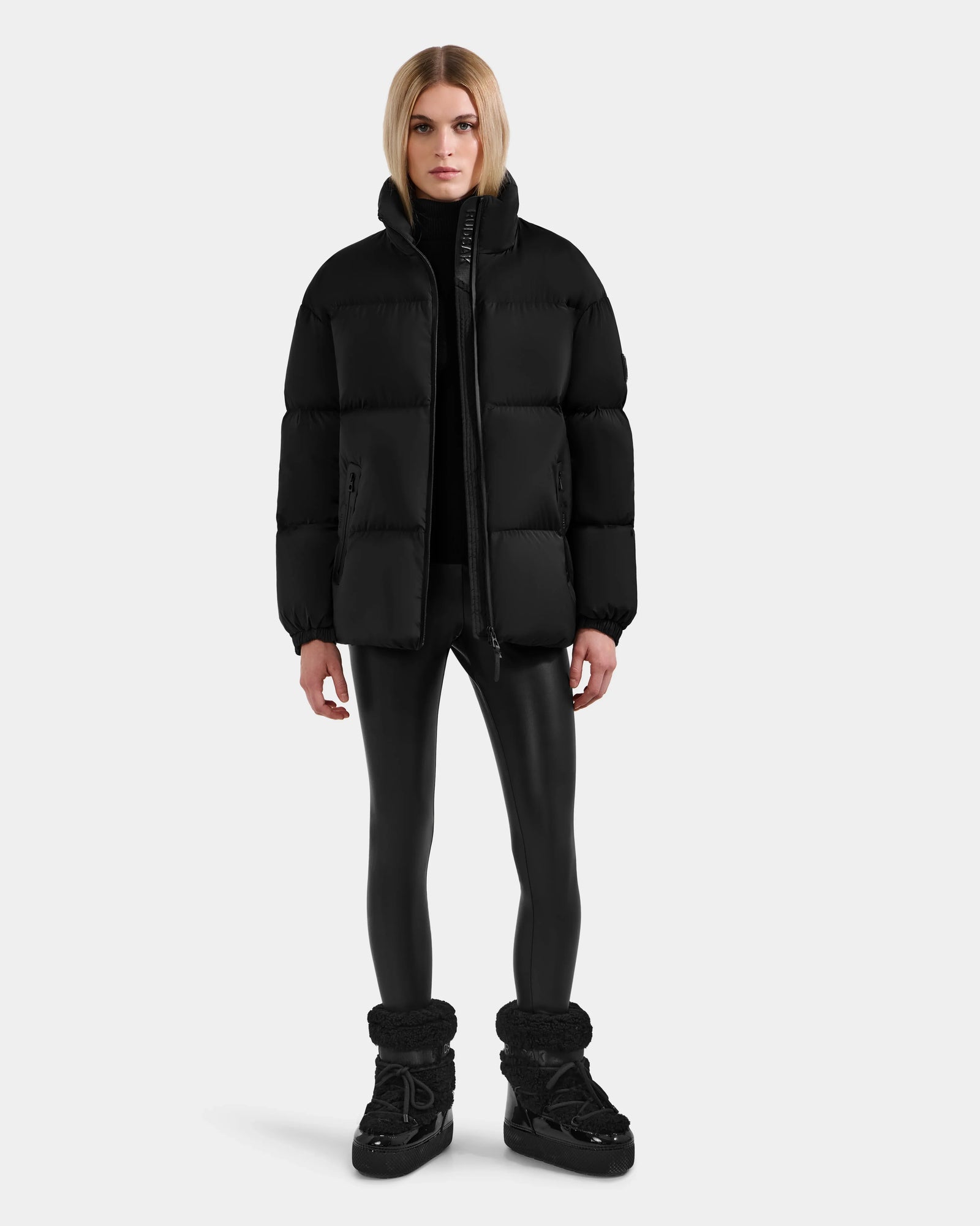 A woman with straight blonde hair faces forward, wearing the Rudsak KARLEE M - 8125681 Black down puffer jacket, black leggings, and winter boots against a plain light gray background.