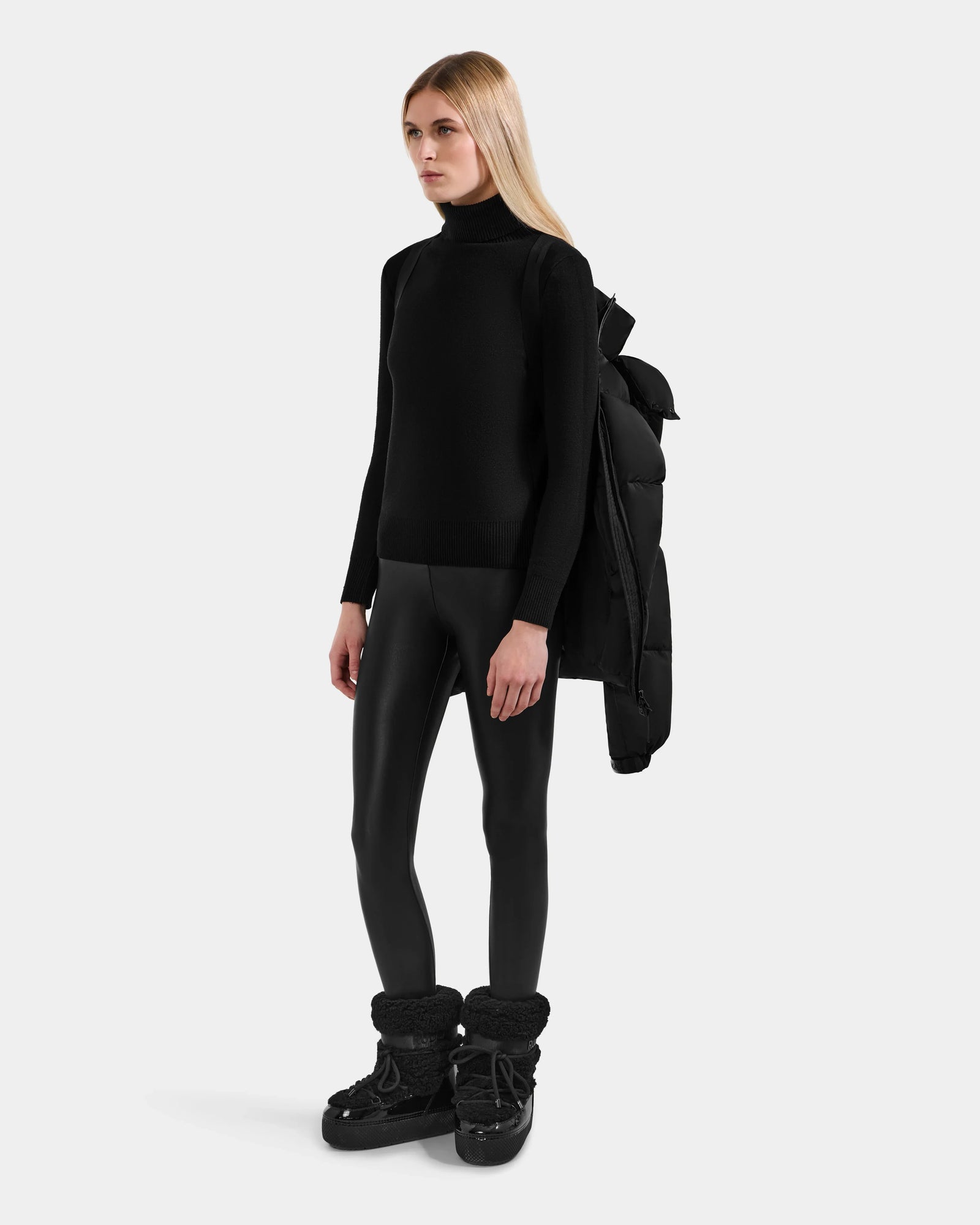A woman with straight blonde hair wears the Rudsak KARLEE M - 8125681 Black down puffer jacket draped over one shoulder, effortlessly showcasing winter warmth against a plain light gray background.