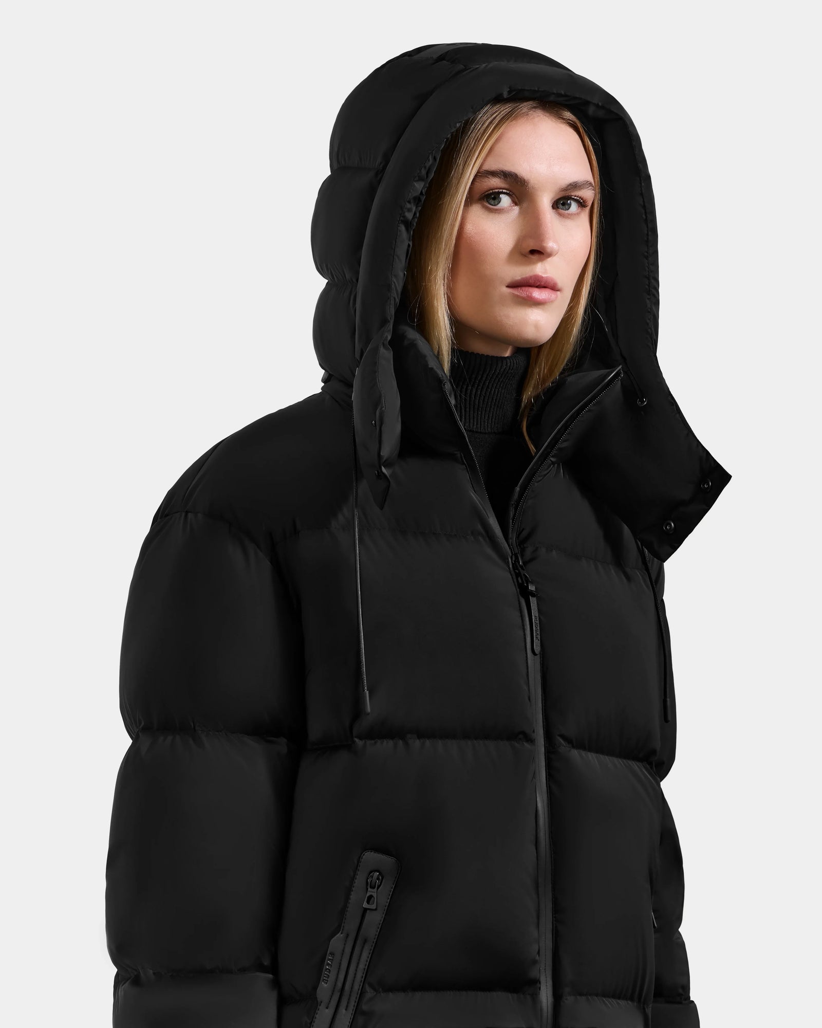 Wearing the Rudsak KARLEE M - 8125681 Black women's down puffer jacket with a hood, she stands against a light gray background, looking slightly to the side—a stylish choice for winter warmth.