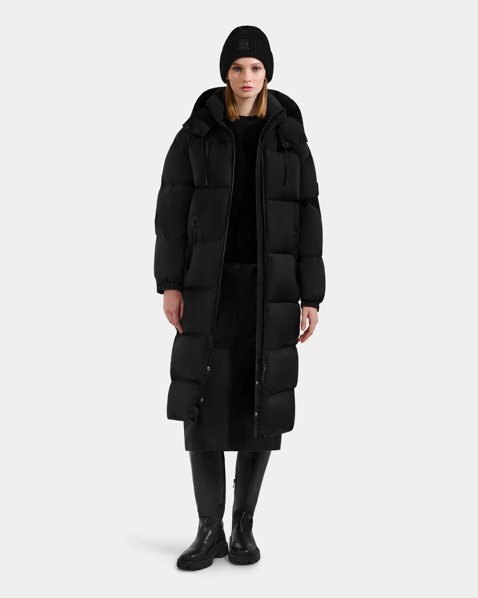 A person models the Rudsak KARLEE XL - 8125683 Black women’s long maxi down puffer jacket with a black beanie, pants, and boots against a plain light gray background—highlighting this essential for extreme cold-weather performance.