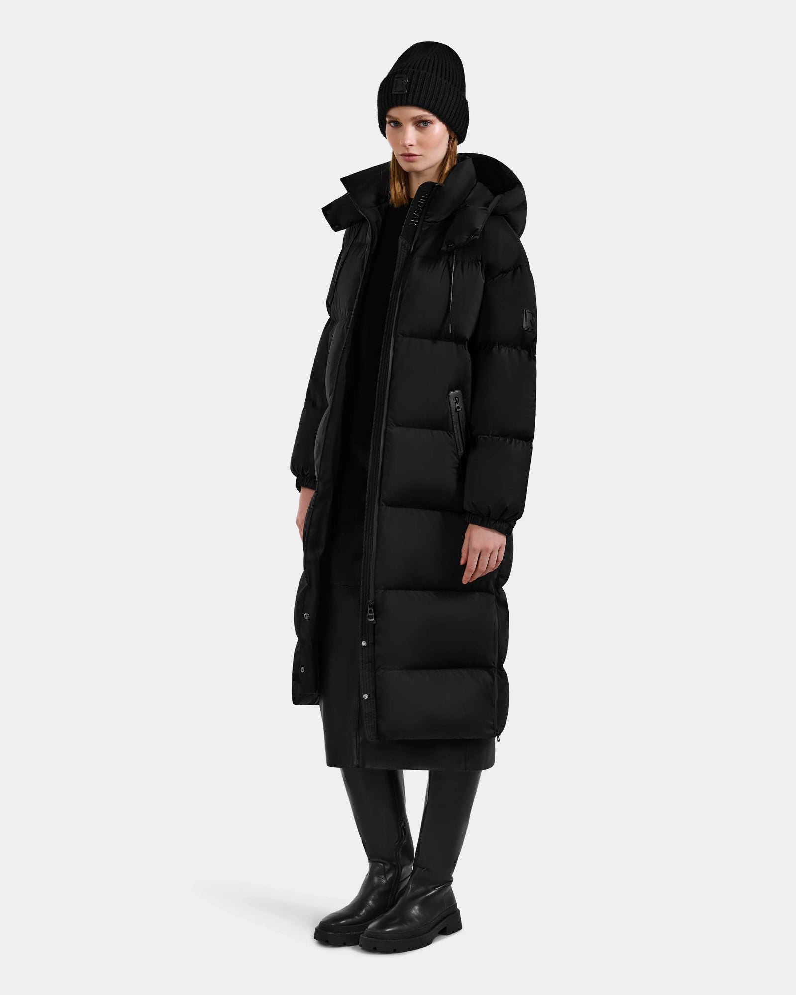 Dressed for extreme cold, a person wears the Rudsak KARLEE XL - 8125683 Black women’s long maxi down puffer jacket, paired with black pants, boots, and a beanie against a simple light backdrop.