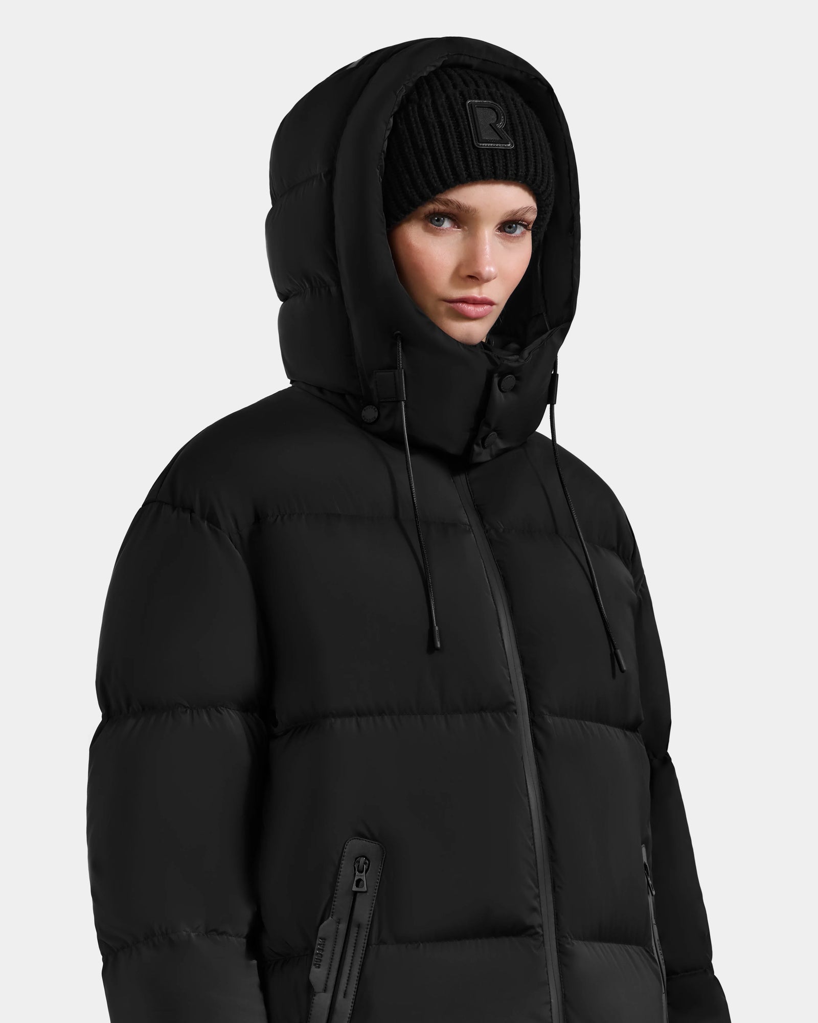Wearing the Rudsak KARLEE XL - 8125683 Black women's maxi down puffer jacket with the hood up and a black knit beanie, she looks forward with a neutral expression against a plain light background—a winter essential.