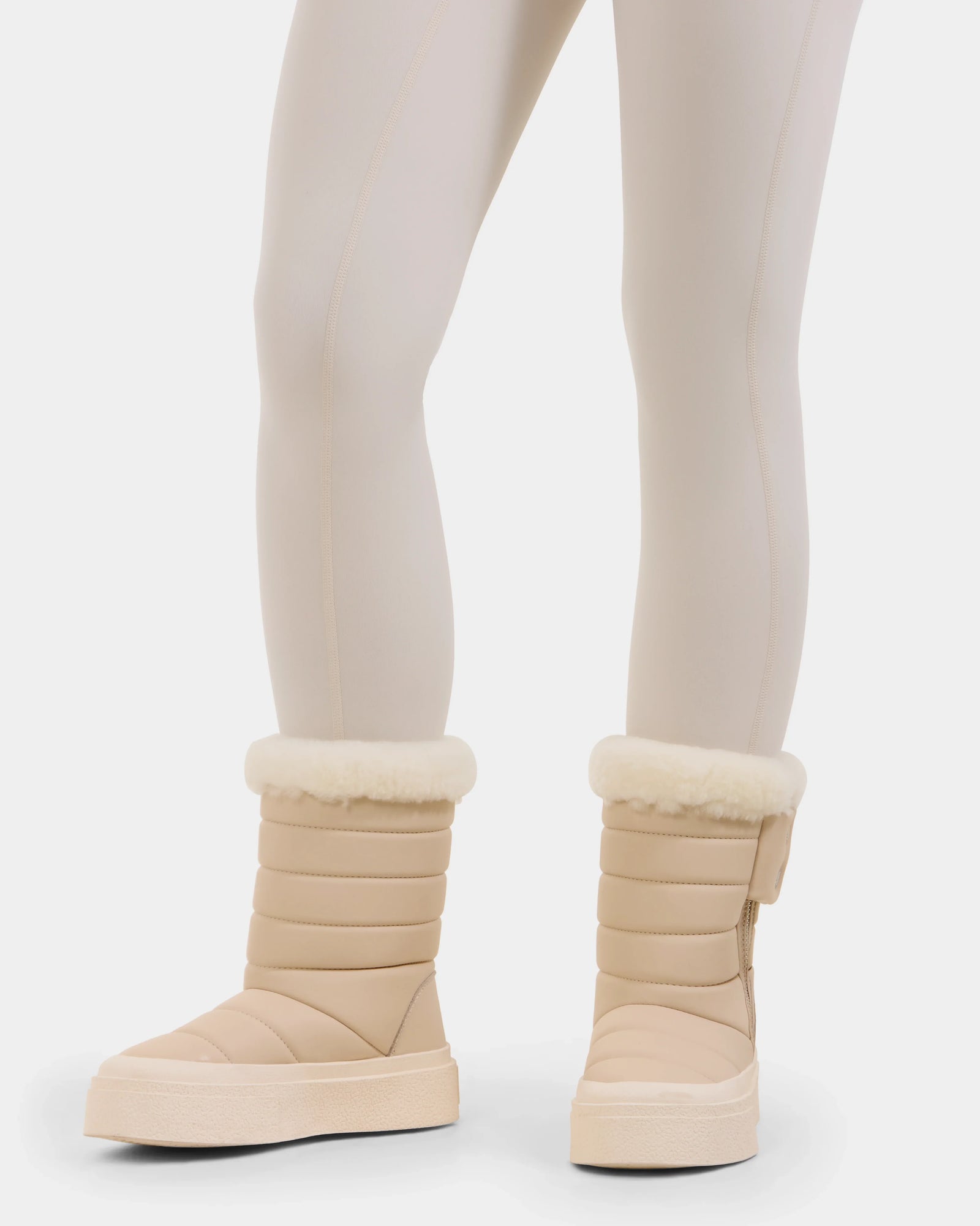 Someone wearing light beige leggings and Rudsak KATHIE - L225516 Vanilla quilted vegan leather winter boots with white fur cuffs stands against a plain white background.