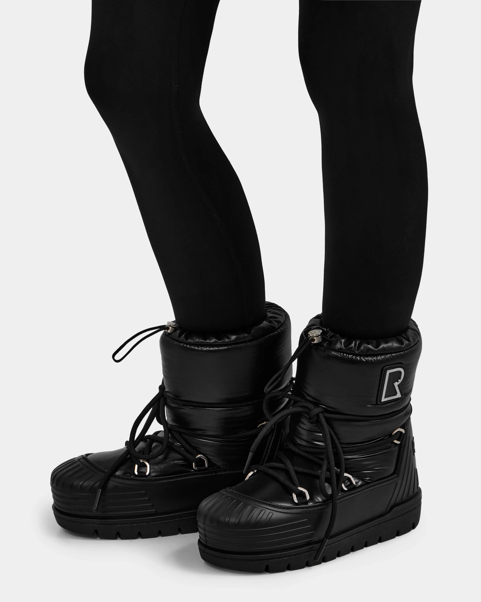 A person wears black leggings and Rudsak KIKA - L224506 Black women's winter boots, featuring chunky rubber soles, laces, an "R" patch, and a quilted, padded texture for added warmth.