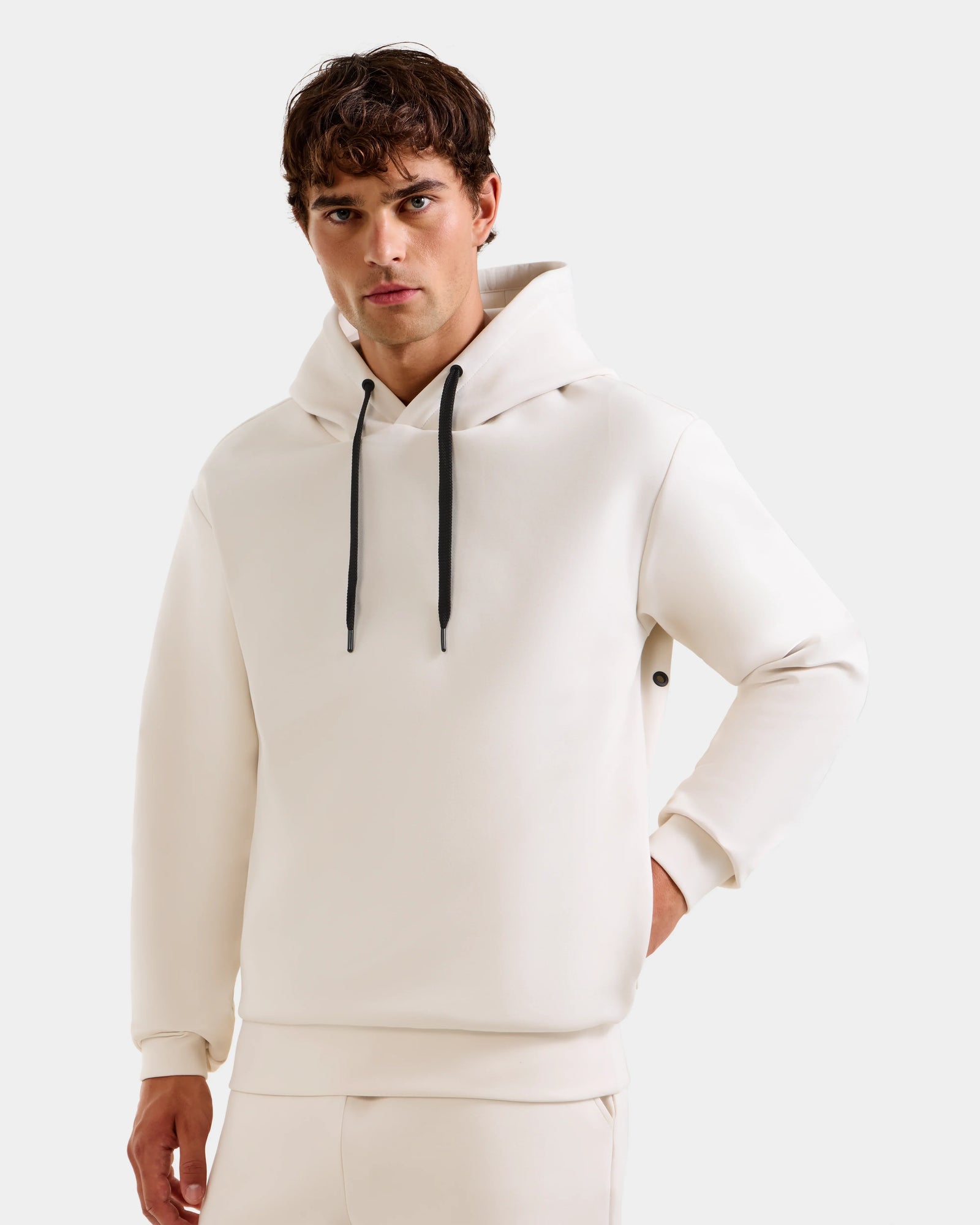 A young man models the Rudsak LEROY - U525509 Vanilla M premium unisex hoodie with black drawstrings and matching pants, standing against a plain light background and looking at the camera, one hand on his hip.