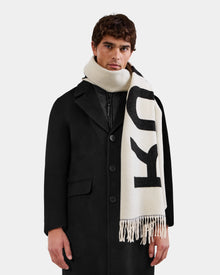 A person wearing the Rudsak LEV - U425506 Black coat and a cream-colored scarf with large black RUDSAK lettering and fringed ends, wrapped around their neck, stands against a plain white background.