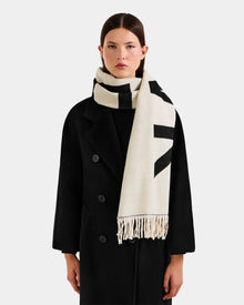 A woman wears the Rudsak LEV - U425506 Black long coat and a thick cream reversible unisex scarf with black patterns and fringed ends, posing against a plain light gray background—an elegant cold-weather look.