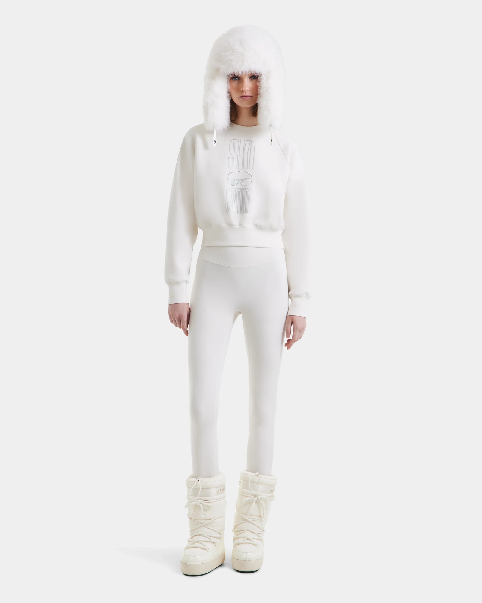 A person stands facing forward in the Rudsak LIDDIE - L525505 Vanilla: an all-white winter look with a furry hat, cropped crewneck sweater, fitted pants, and puffy high snow boots, set against a plain white background.
