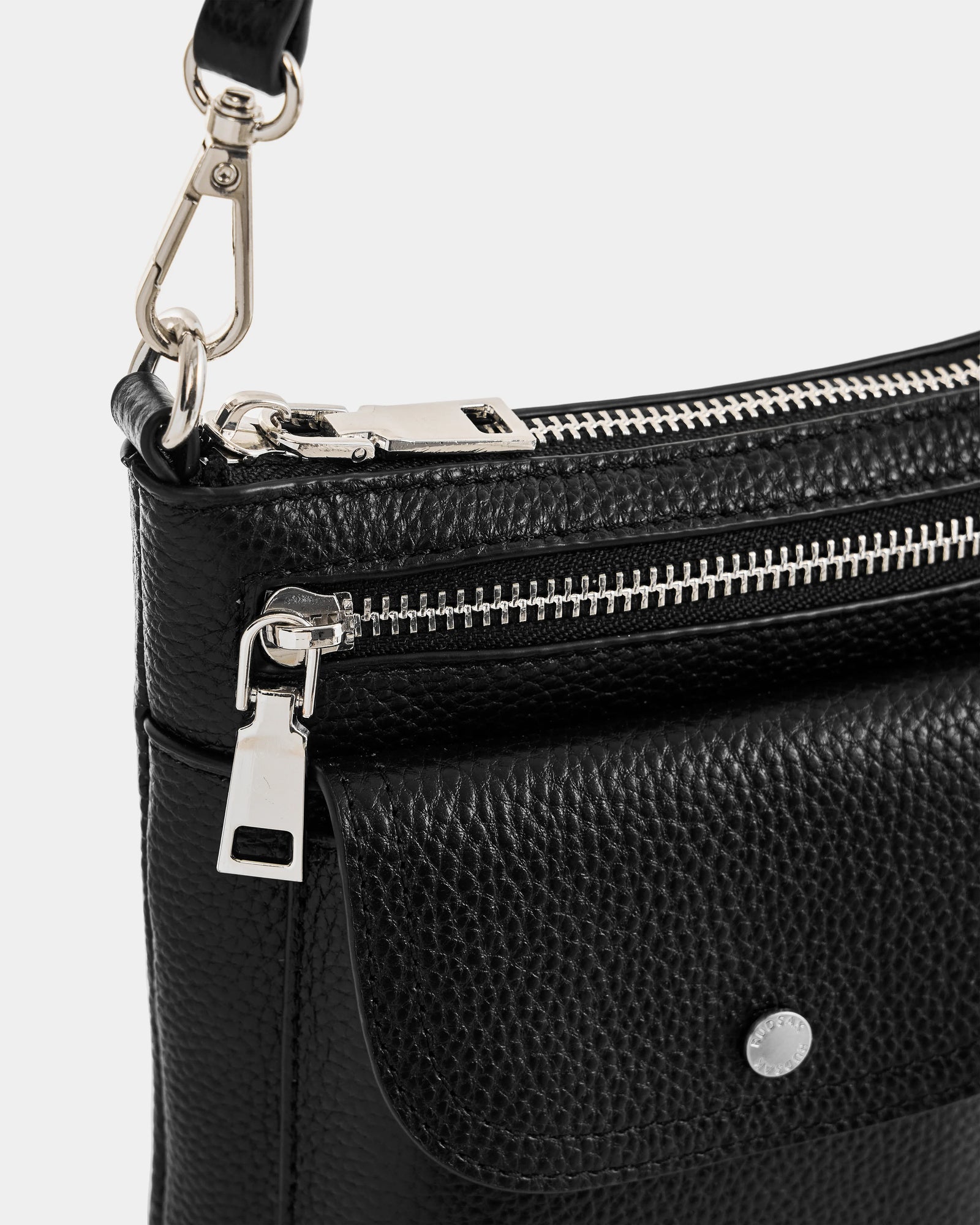 Close-up of the Rudsak LILA - L325513 Black women’s crossbody bag in black pebbled leather with a front flap pocket, silver zipper, metal clasp fastenings, adjustable strap, and visible hardware details.