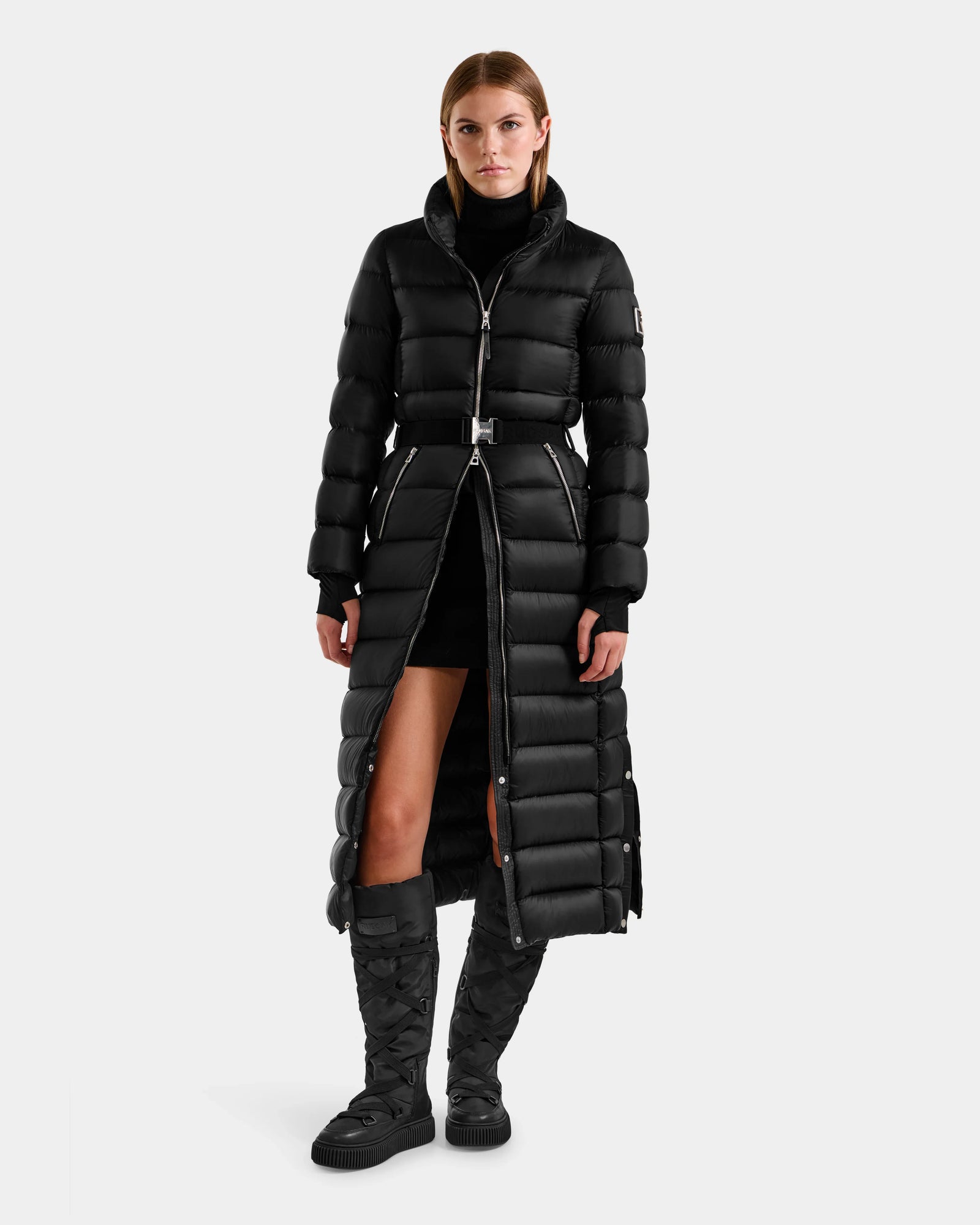 A woman wearing the Rudsak LILITH L - L125528 Black quilted puffer coat stands against a plain light gray background, styled with black knee-high quilted boots and a black dress.