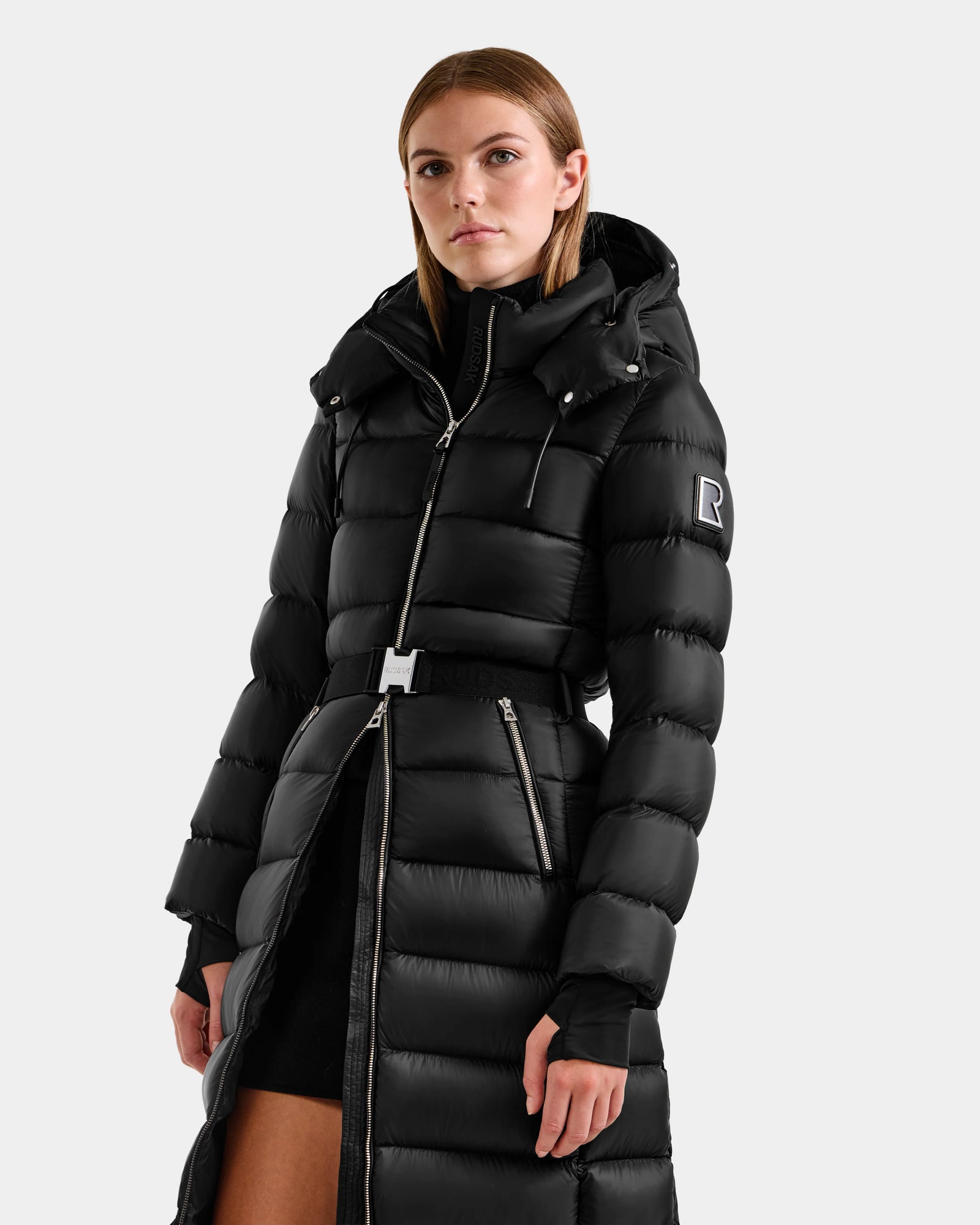 A woman stands against a plain background in the LILITH L - L125528 Black puffer coat by Rudsak, featuring a hood, zipper, belted waist, and a sleeve patch with the letter R.