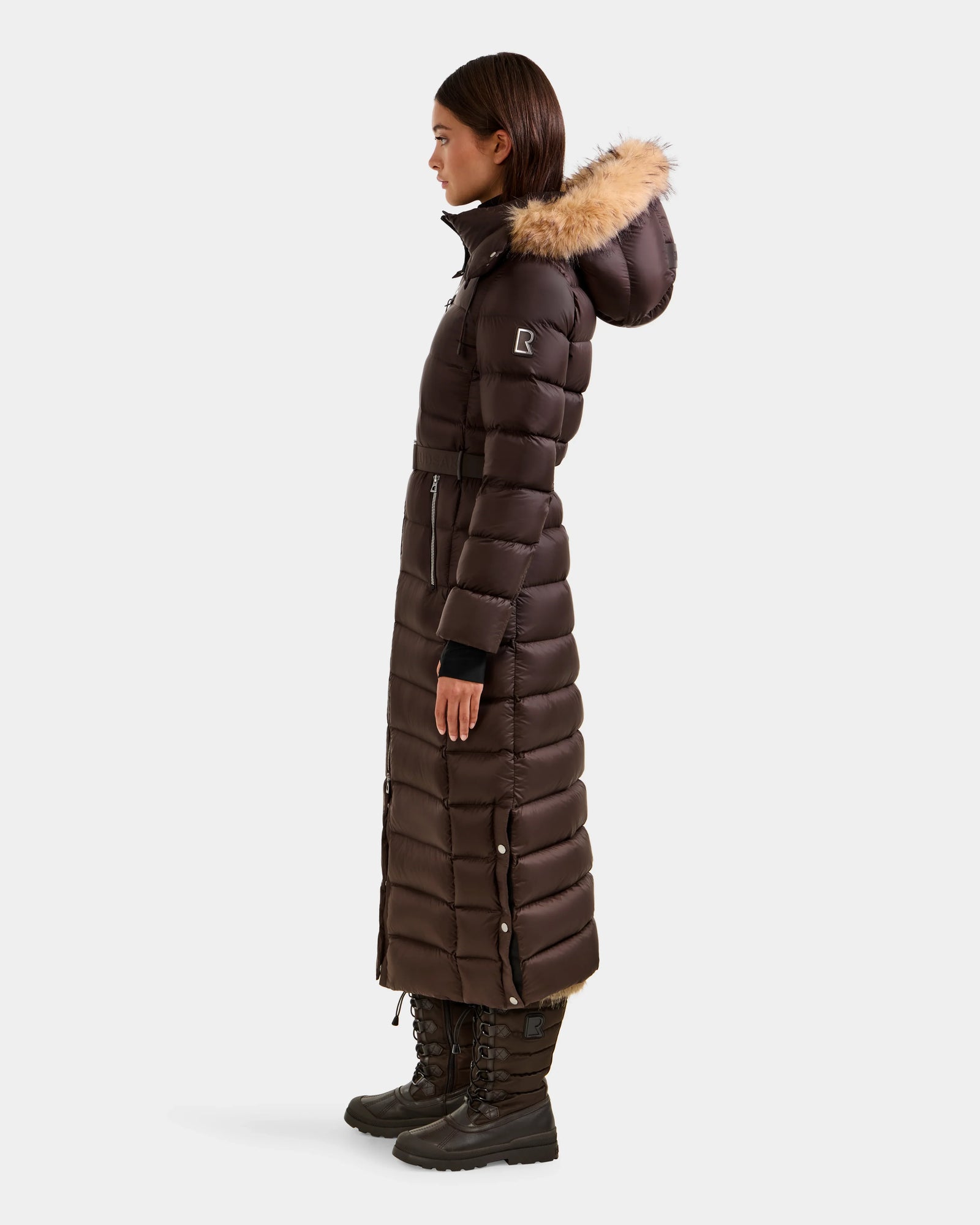 A woman stands in profile wearing the Rudsak LILITH L - L125528 Espresso, a dark brown maxi-length puffer with RDS-certified down, fur-trimmed hood, removable belt, and quilted design. She pairs it with black boots against a plain white background.