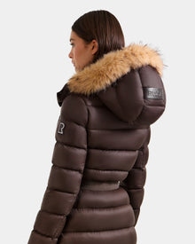 A woman wears the Rudsak LILITH L - L125528 Espresso maxi down puffer with a fur-trimmed hood, RDS-certified down, a black arm patch, and “RUDSAK” on the hood, shown from the side and back.