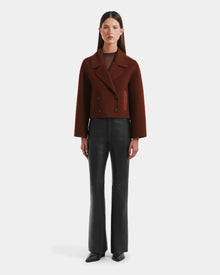 A woman faces forward against a white background wearing the Rudsak MARTINEZ - 8125580 Rust cropped double-breasted blazer, black vegan leather pants, and black pointed-toe shoes.