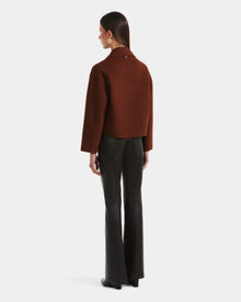 A woman with straight brown hair faces away, wearing the Rudsak MARTINEZ - 8125580 Rust short coat and black flared pants against a plain white background.