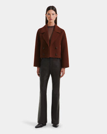 A woman faces forward against a white background wearing the Rudsak MARTINEZ - 8125580 Rust cropped double-breasted women’s short coat over a sheer brown top, paired with black vegan leather flared pants and black shoes.