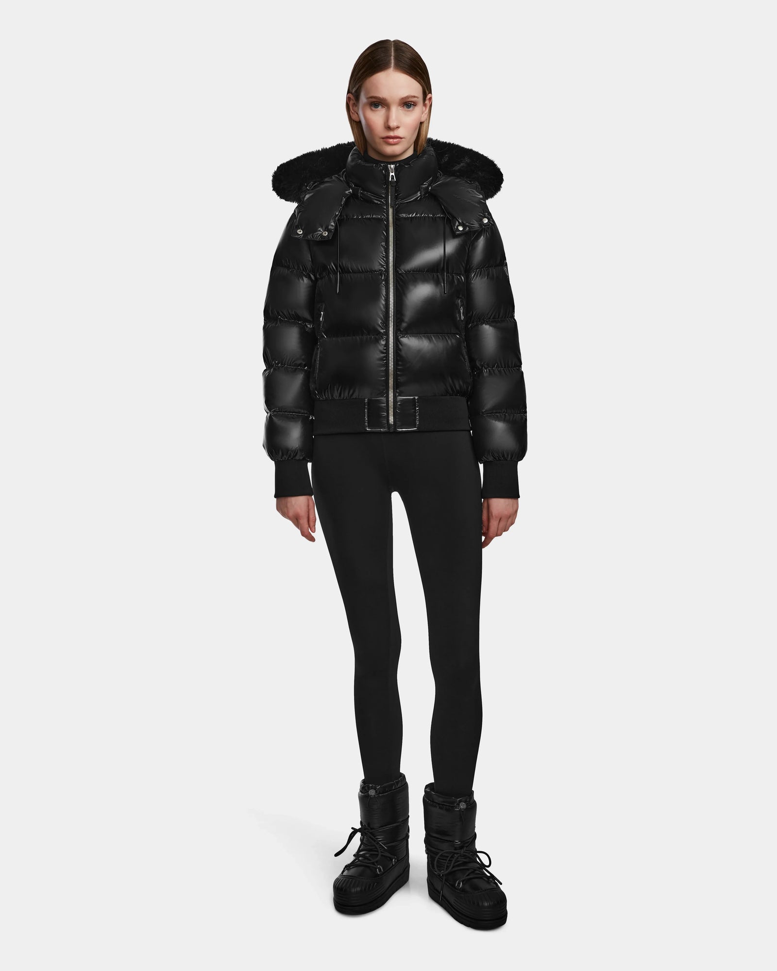 A woman stands facing forward wearing the Rudsak MAUDE Q-S - L124553 Black RDS-certified down jacket with a hood, paired with black leggings and winter boots against a plain white background.