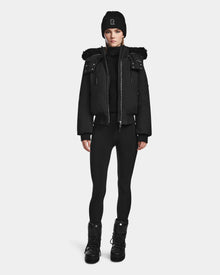 A person stands facing forward in a black Rudsak MAUDE - L124522 jacket with fur trim, paired with a black beanie, turtleneck, leggings, and lace-up boots against a plain white background.