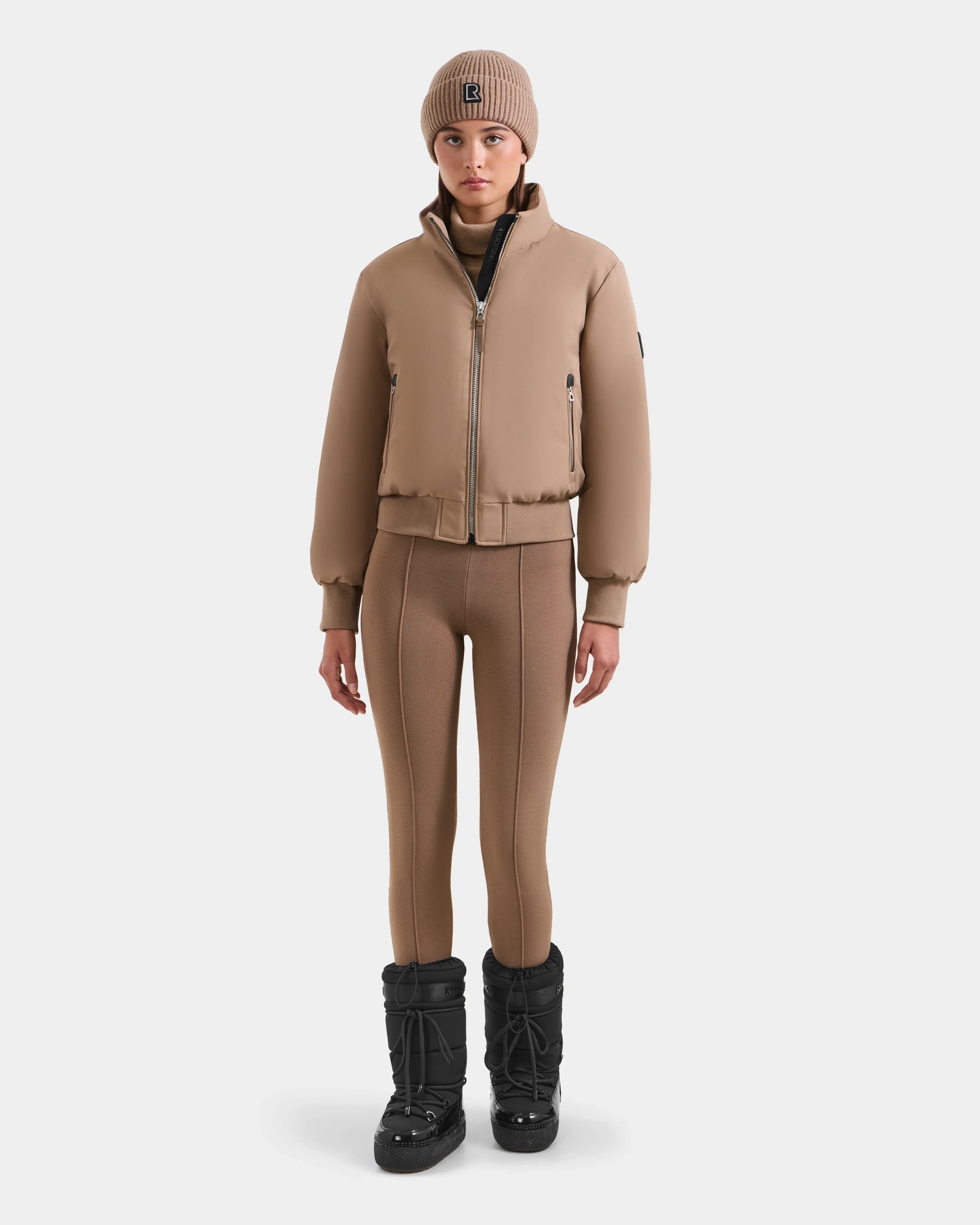 A person stands facing forward against a white background, wearing the Rudsak MAUDE - L124522 Sand women's down bomber, paired with a matching tan knit beanie, tan leggings, and black winter boots.