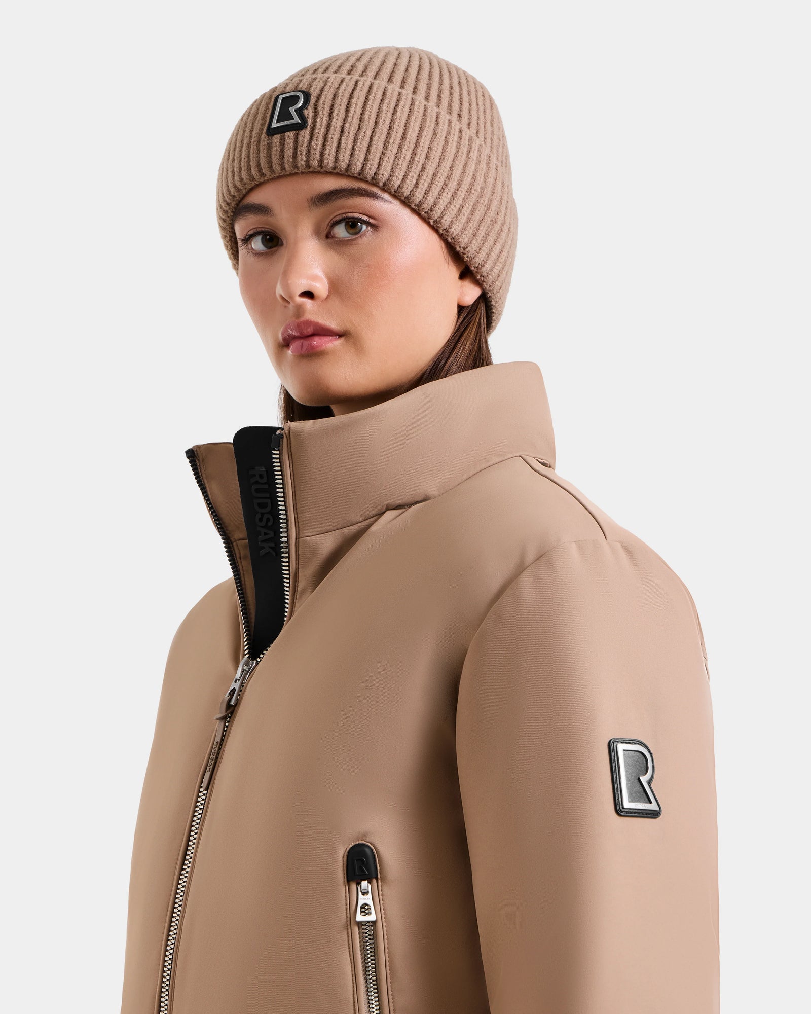 A person wears the Rudsak MAUDE - L124522 Sand beanie and matching winter jacket with black R logo patches, looking slightly to the side against a plain light background.