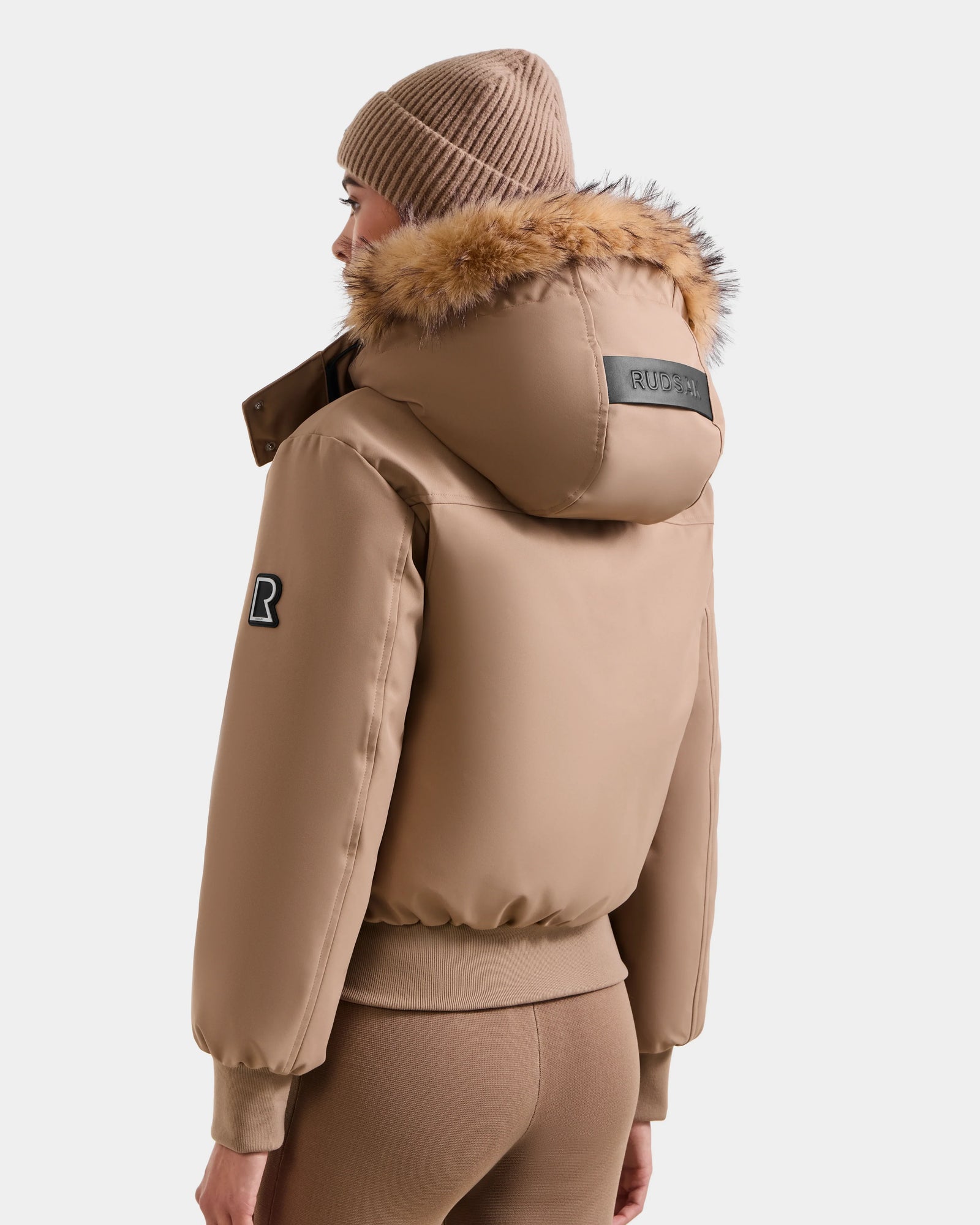 A person models the Rudsak MAUDE - L124522 Sand jacket, featuring premium down insulation, a fur-trimmed hood, and a black patch, paired with a matching knit beanie and fitted pants, standing against a white background.