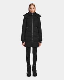 A woman faces forward in the Rudsak MIRIAM - L123544 Black down puffer coat with high collar and hood, paired with black leggings and boots, against a plain white background.