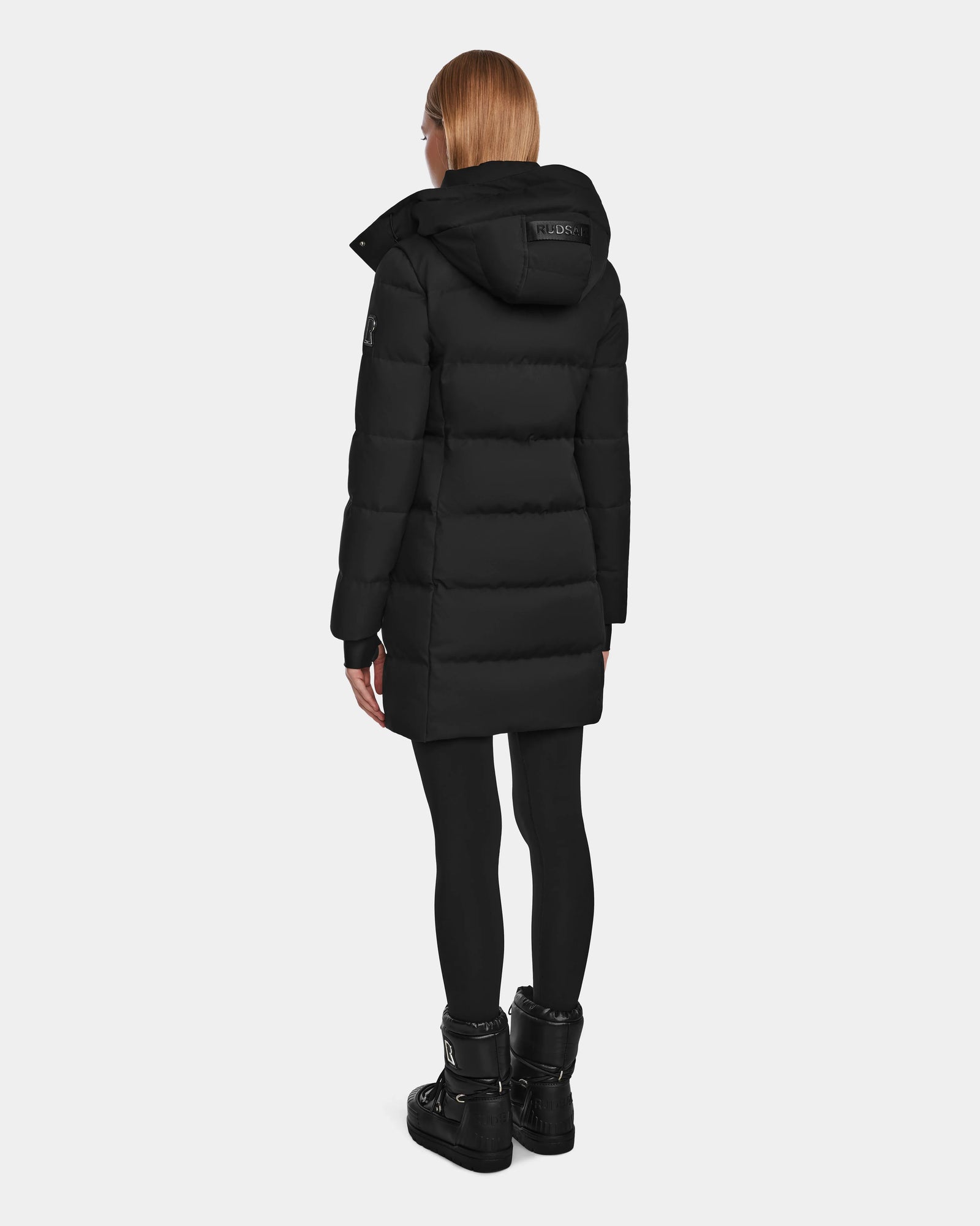 A person with straight, shoulder-length hair is seen from the back wearing the Rudsak MIRIAM - L123544 Black down puffer with a hood, paired with black leggings and boots, standing against a plain light background.