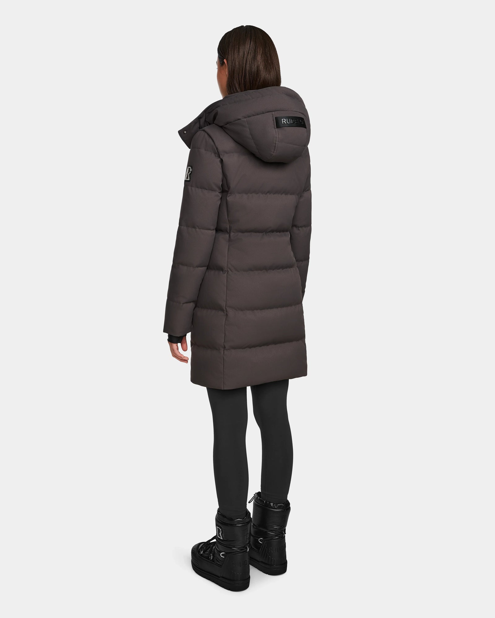 A person wearing the Rudsak MIRIAM - L123544 Grey womens down puffer coat with a hood, paired with black leggings and black boots, stands facing away from the camera against a plain white background.