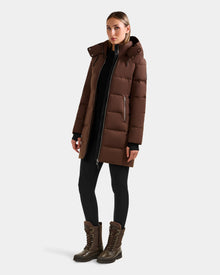A woman faces forward wearing the Rudsak MIRIAM - L123544 Mocha, a women’s down puffer coat with a weather-resistant shell, paired with black leggings and brown lace-up boots against a plain light background.