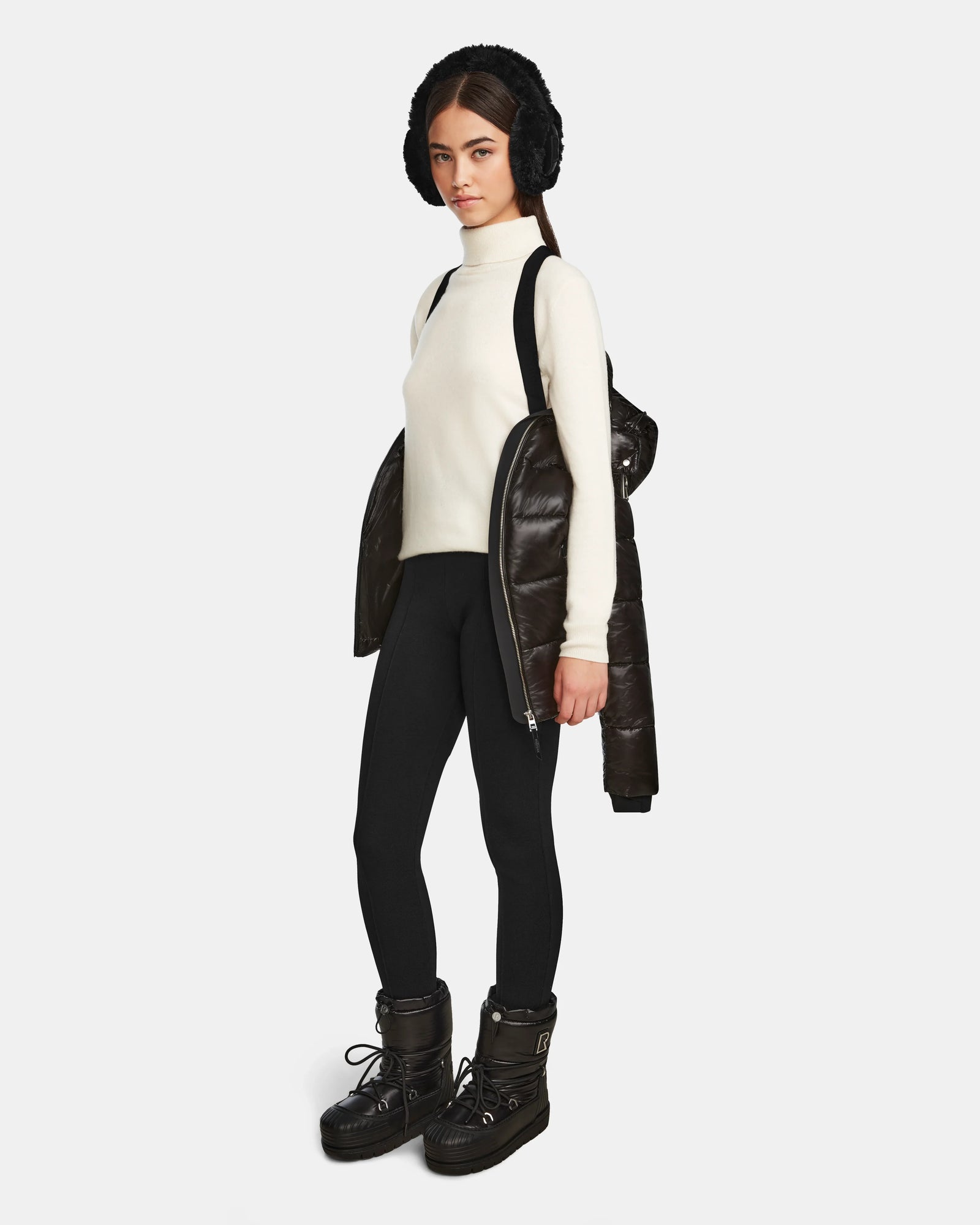A woman faces forward in a white turtleneck, black leggings, winter boots, and a Rudsak MIRRY S - L123545 Black quilted down coat over her shoulders, with a black furry hat. The background is plain white.