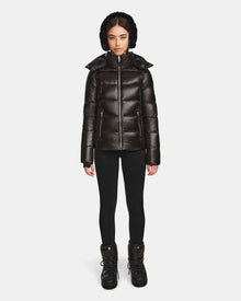 A woman faces forward in a shiny black Rudsak MIRRY S - L123545 winter coat with a hood, paired with black leggings and winter boots. The background is plain white.