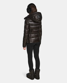 A person stands facing away wearing black earmuffs, black leggings, black winter boots, and a shiny Rudsak MIRRY S - L123545 Black womens quilted down coat with hood against a plain white background.