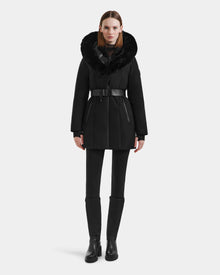 A woman faces forward against a white background, wearing the Rudsak MODA M - L125555 Black down puffer coat with a vegan leather belt and faux fur hood, paired with black leggings and boots.