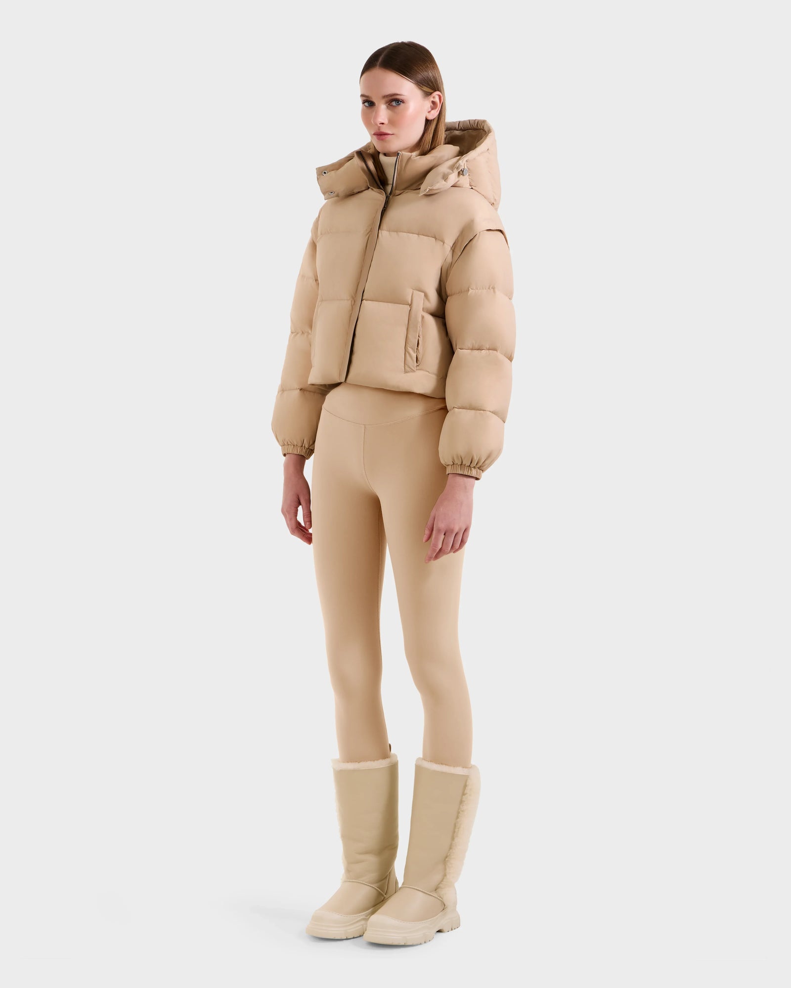 A woman faces forward wearing the Rudsak OPRAH - L125546 Vanilla cropped down puffer jacket with removable sleeves, paired with matching beige leggings and knee-high winter boots, set against a plain white background.