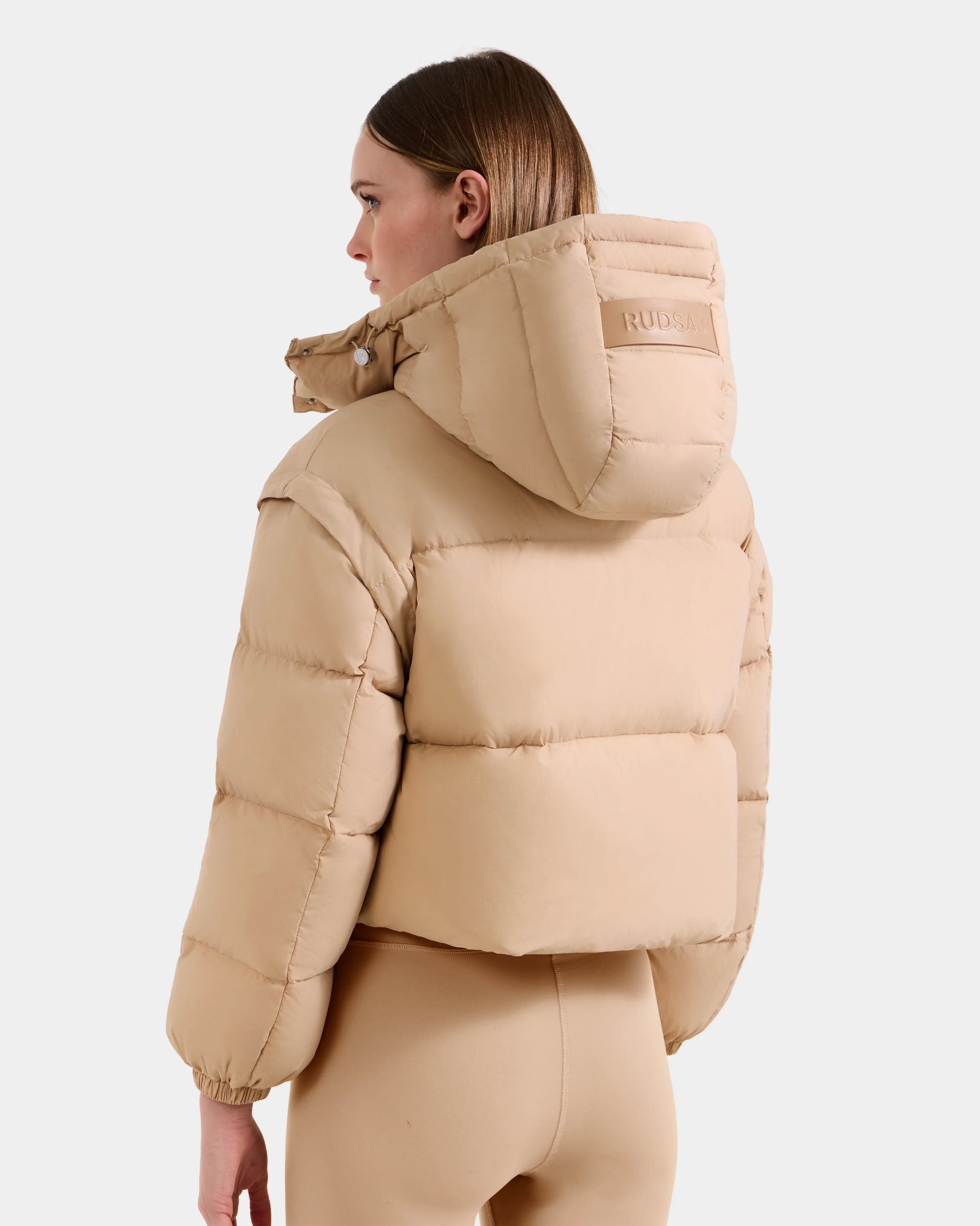 A woman with straight brown hair, facing away from the camera against a plain light background, wears the Rudsak OPRAH - L125546 Vanilla women’s cropped down puffer with hood and matching leggings.