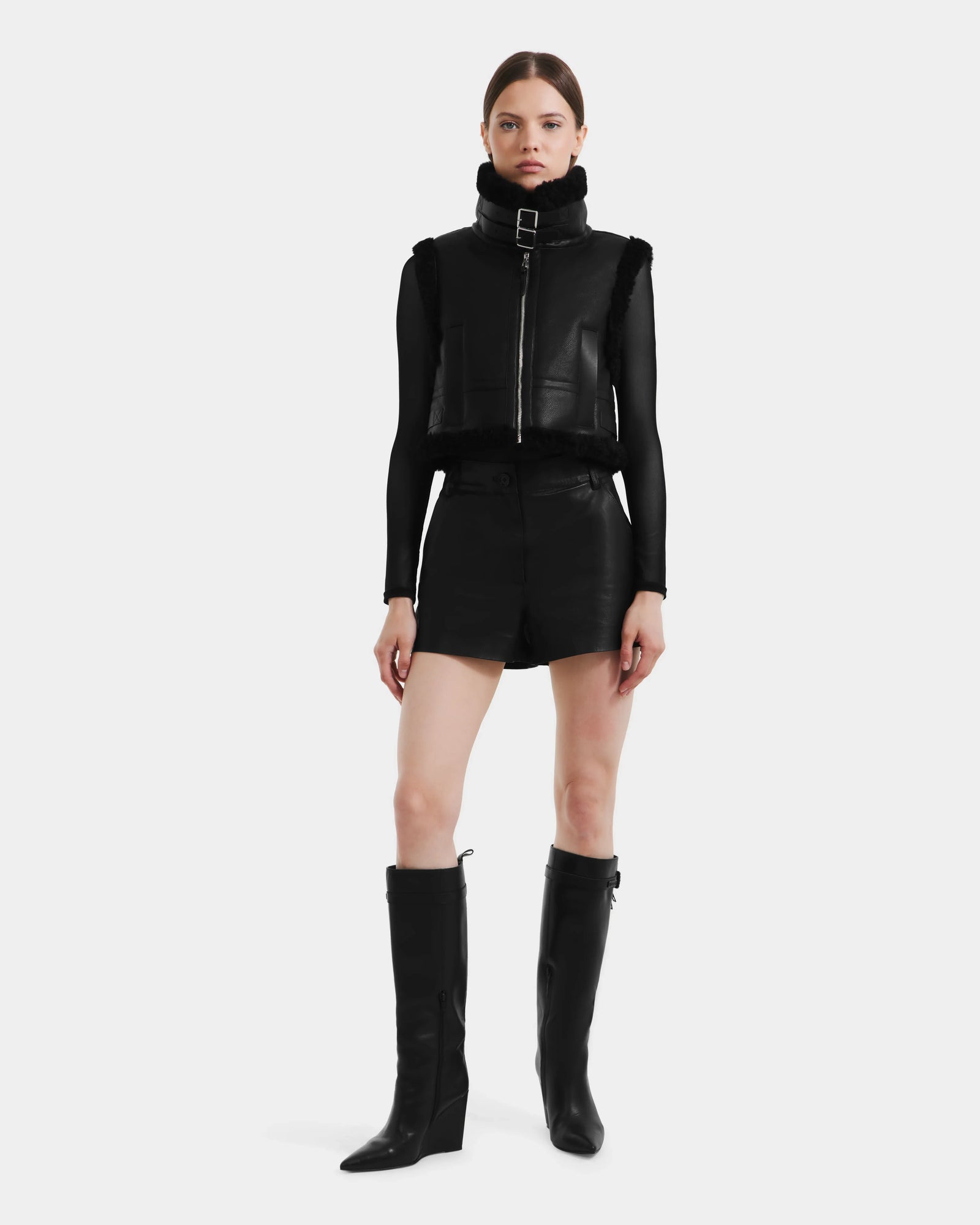A woman with straight, pulled-back hair and a neutral expression wears the Rudsak PAISLEY - L125516 Black women's cropped vegan leather vest, styled with black shorts, knee-high boots, and a sheer long-sleeve black top.