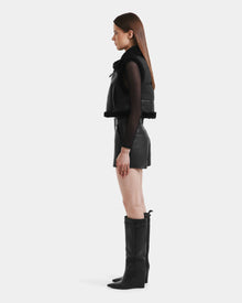 A woman stands in profile against a white background, wearing the Rudsak PAISLEY - L125516 Black faux shearling vest over a black sheer long-sleeve top, paired with black leather shorts and knee-high black boots.