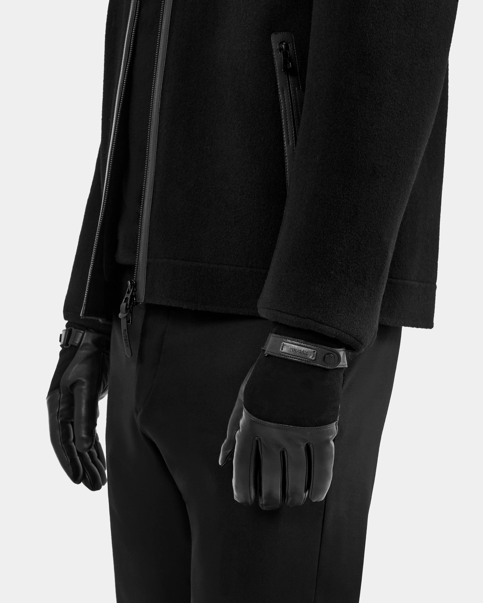 A person dressed in all black, wearing Rudsak’s PARKER - M425502 Black zipped jacket, matching pants, and men’s leather gloves with a Velcro strap and wool-cashmere lining stands against a plain light background.