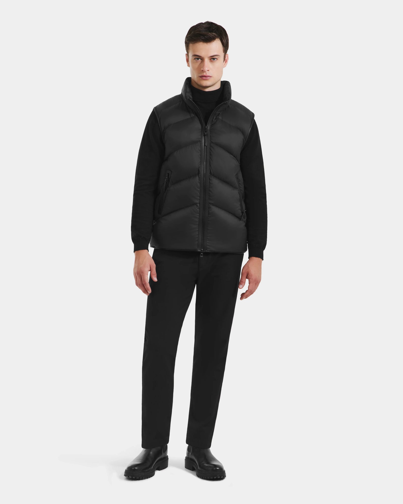 A man faces forward in a Rudsak RICHARDSON - M125527 Black men's hooded vest over a black turtleneck, pants, and shoes against a white background. The vest provides water-repellent protection and lightweight warmth for comfort.