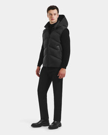 A man faces slightly left, wearing the Rudsak RICHARDSON - M125527 Black men’s hooded vest over a black turtleneck, with black pants and boots, set against a plain white background.