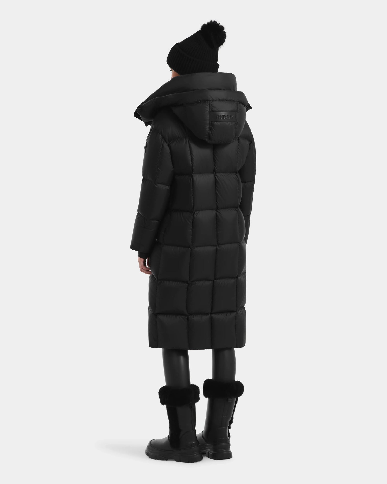 A person wears the Rudsak SASHA - 8125686 Black women’s long down puffer coat with removable hood, paired with black leggings, fur-trimmed boots, and a black beanie, standing against a plain light gray background.